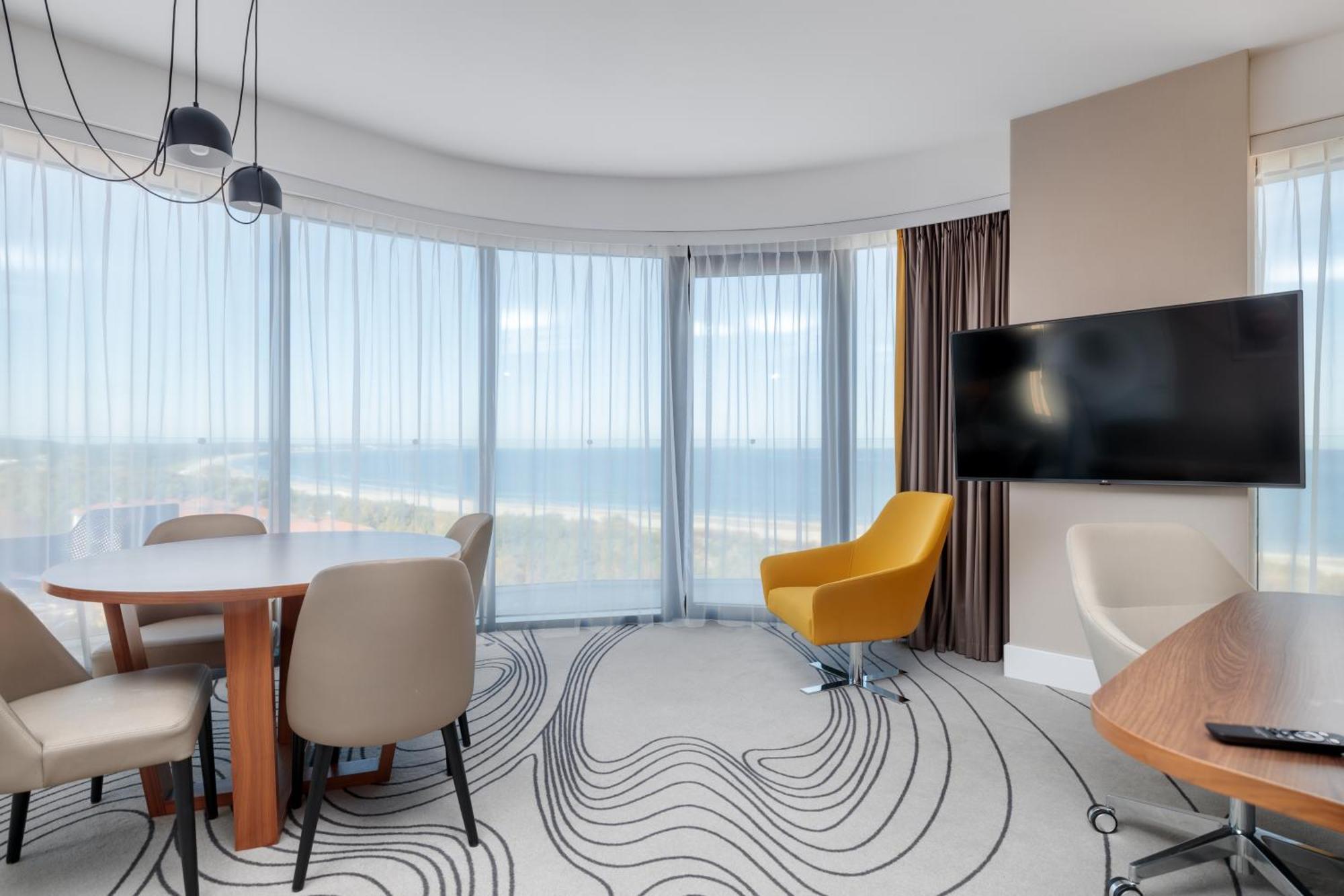 Radisson Blu Resort Swinoujscie Kültér fotó The image shows a modern hotel room or suite with a bright, airy atmosphere. There are large windows that allow natural light to fill the space, providing a view of the ocean. The decor includes a round dining table with several chairs, and a stylish