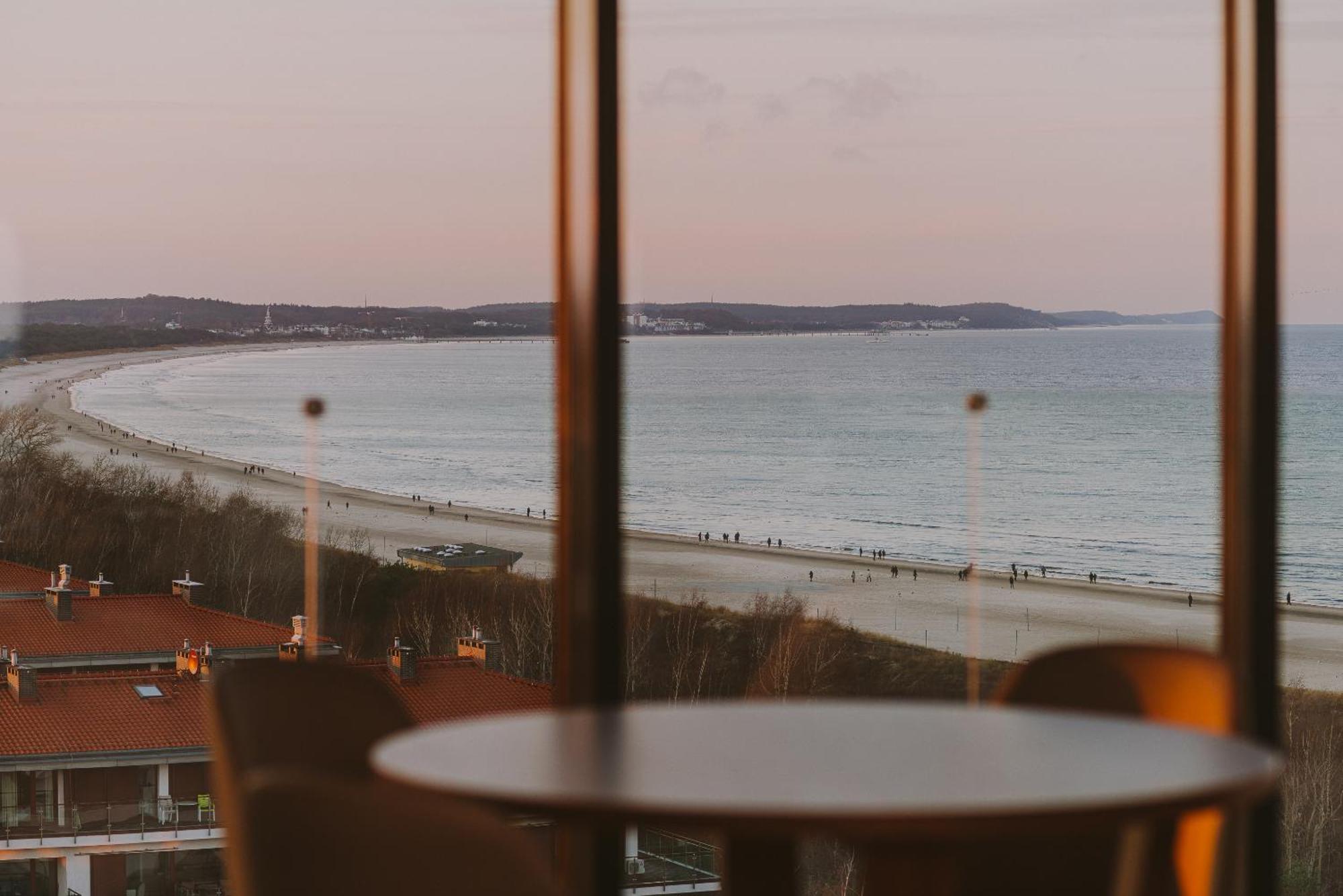 Radisson Blu Resort Swinoujscie Kültér fotó The photo shows a picturesque view of a beach scene from indoors. In the foreground, there is a round table with some chairs around it, suggesting a cozy interior space. Through a large window, you can see the beach with gentle waves hitting the shor