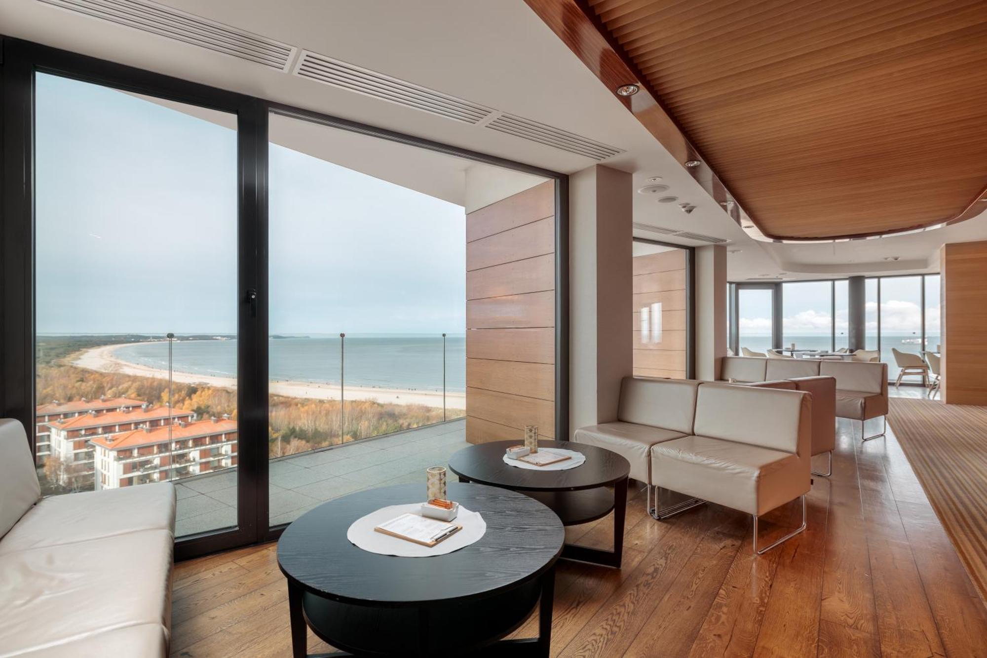 Radisson Blu Resort Swinoujscie Kültér fotó The photo shows a modern, elegantly designed interior space featuring large windows that offer a panoramic view of the ocean and coastline. The flooring is wooden, providing a warm atmosphere. There are two round black tables in the foreground with m