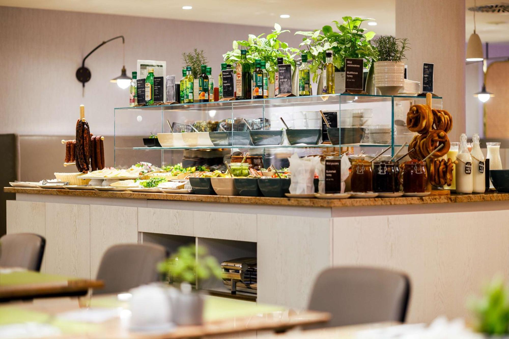 Radisson Blu Resort Swinoujscie Kültér fotó The photo shows a well-organized buffet display in a restaurant setting. There is a glass counter filled with various dishes. On the upper shelf, there are several bottles of oil and herbs, indicating a focus on fresh and healthy ingredients. Below t