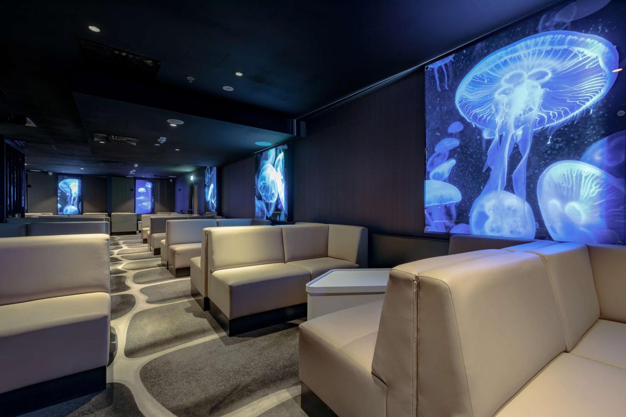 Radisson Blu Resort Swinoujscie Kültér fotó The photo shows a modern lounge space with a sleek design. The area features light-colored, upholstered seating arranged in an open layout. The walls are decorated with large, vibrant images of jellyfish illuminated in blue, creating a calming underw