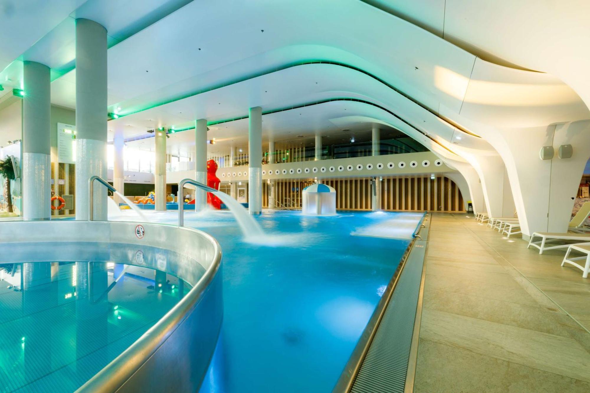 Radisson Blu Resort Swinoujscie Kültér fotó The photo shows a modern indoor swimming pool area with an elegant design. The pool features curved edges and is filled with clear, blue water. There are various amenities, such as jets for relaxation and possibly seating areas along the edges. The l