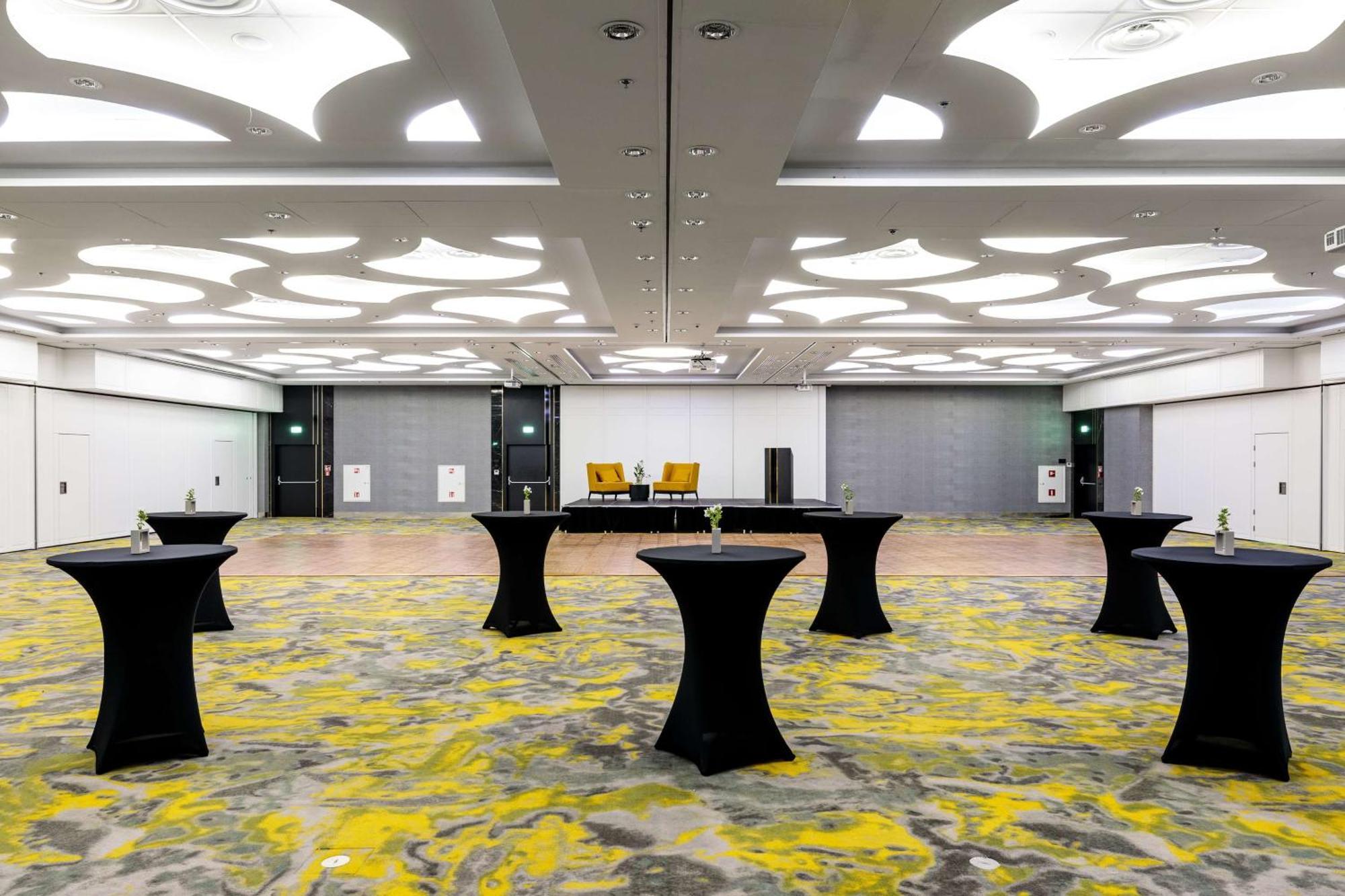 Radisson Blu Resort Swinoujscie Kültér fotó The photo shows a spacious interior room set up for an event. The flooring features a patterned design in gray with yellow accents. There are several black tall tables scattered throughout the area, and in the background, there are two large yellow a