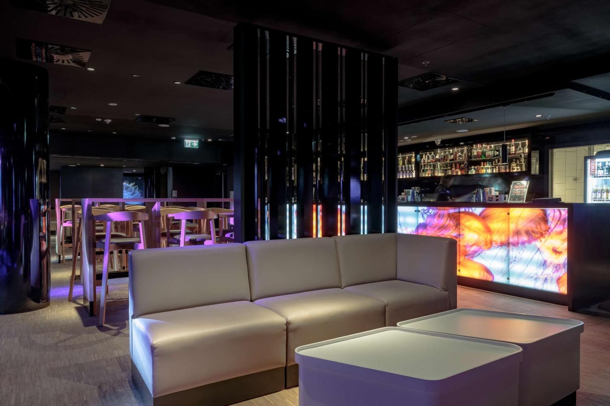 Radisson Blu Resort Swinoujscie Kültér fotó The photo depicts a modern interior space, possibly a lounge or bar. There are sleek, minimalistic furniture pieces, including a large, curved sofa and contemporary coffee tables. The lighting is dim, with a blend of colorful ambient lights illuminat