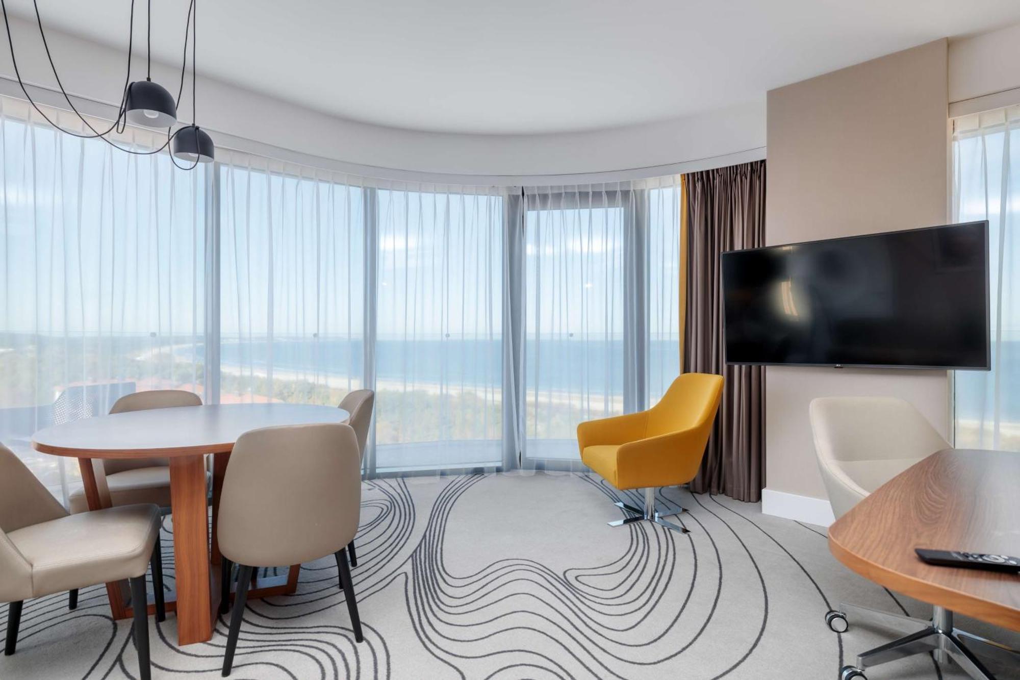Radisson Blu Resort Swinoujscie Kültér fotó The photo shows a modern, stylish interior of a room with large windows that provide a panoramic view. The space features a round dining table surrounded by chairs, including a distinct yellow armchair. There is a flat-screen television mounted on th