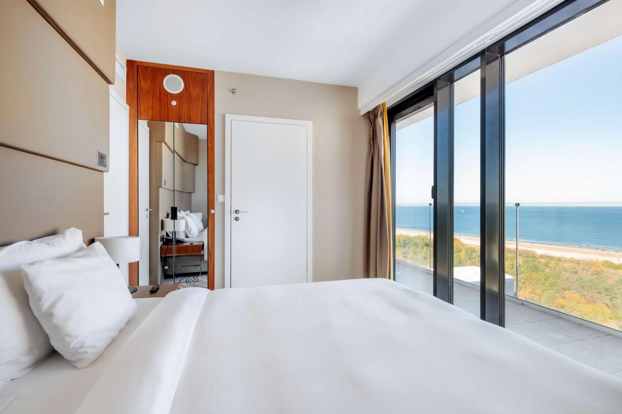 Radisson Blu Resort Swinoujscie Kültér fotó The photo shows a modern hotel room with a bright and airy atmosphere. There is a large bed with white bedding positioned near a glass wall that offers a view of the ocean. The room features neutral-colored walls and wooden accents, enhancing its con