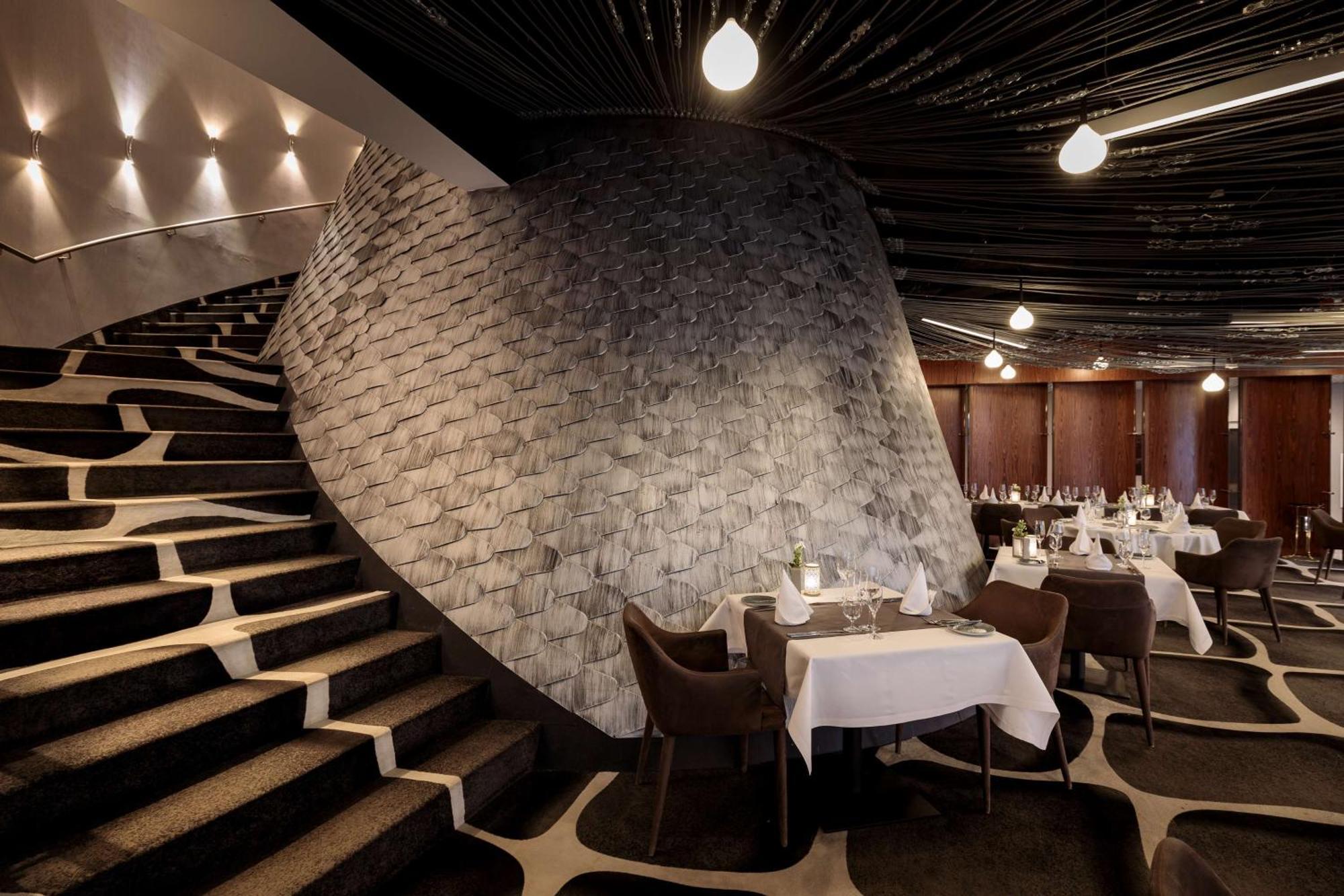Radisson Blu Resort Swinoujscie Kültér fotó The photo shows the interior of a stylish restaurant. In the foreground, there are elegantly set tables with white tablecloths and dark chairs. A unique, curving stone wall adds a distinct architectural feature, and a grand staircase with a textured 