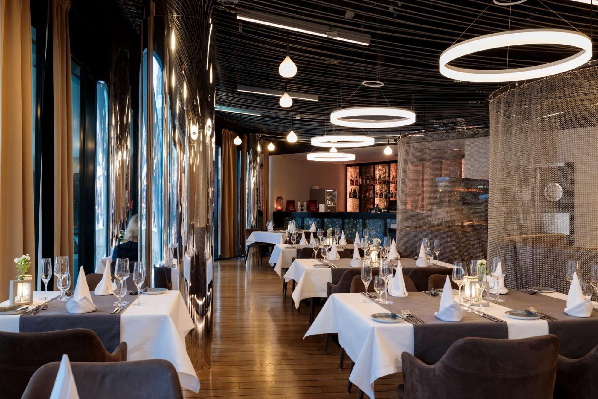 Radisson Blu Resort Swinoujscie Kültér fotó The photo shows the interior of a modern restaurant. It features elegantly set tables with white tablecloths and neatly arranged glassware. The design includes a sleek, contemporary aesthetic with dark walls and interesting lighting, including circul