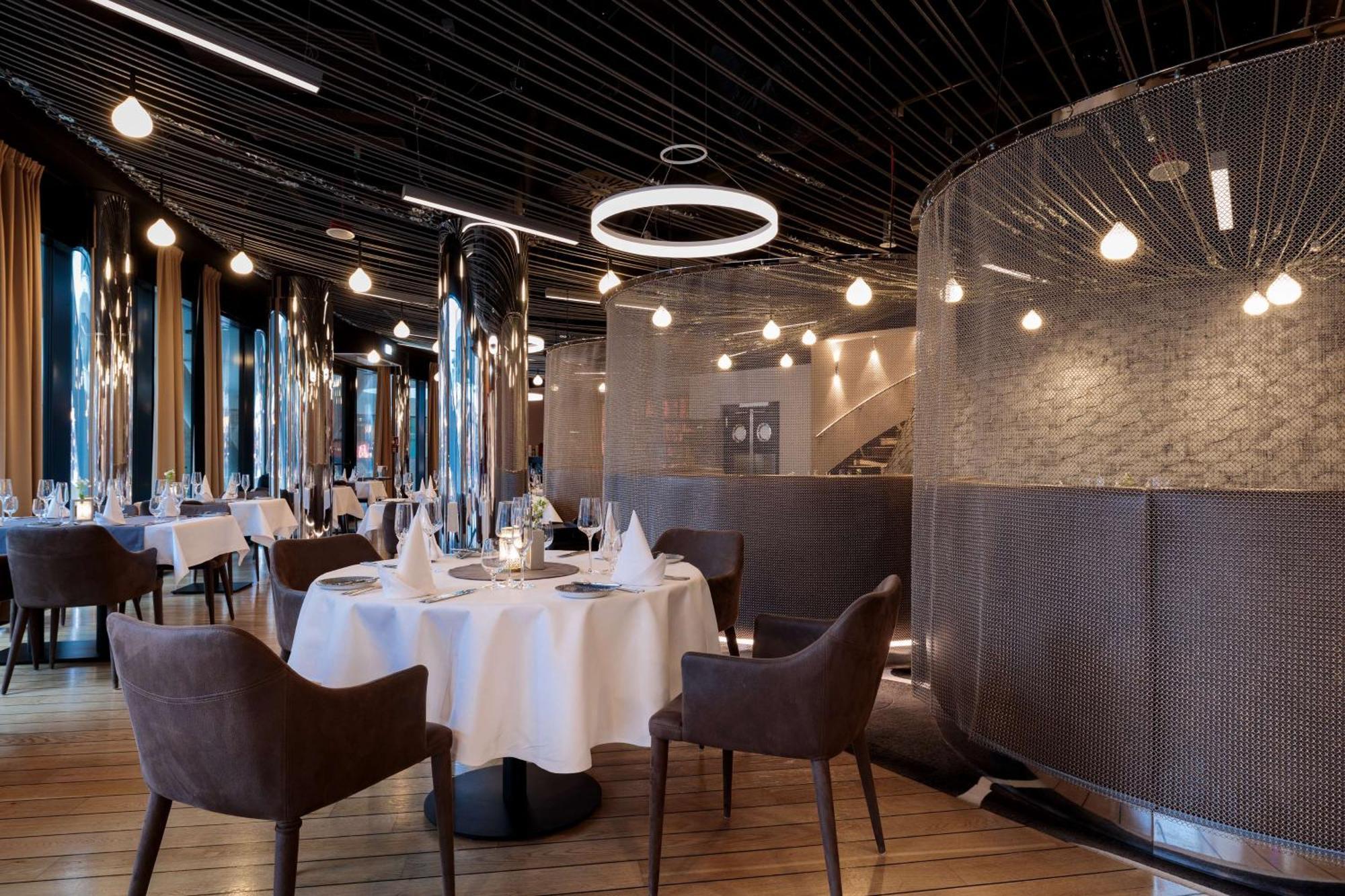 Radisson Blu Resort Swinoujscie Kültér fotó The image shows a modern restaurant interior that features a stylish design. There are several round tables with white tablecloths. The seating consists of brown, upholstered chairs. The space is illuminated by various hanging lights, including a dis