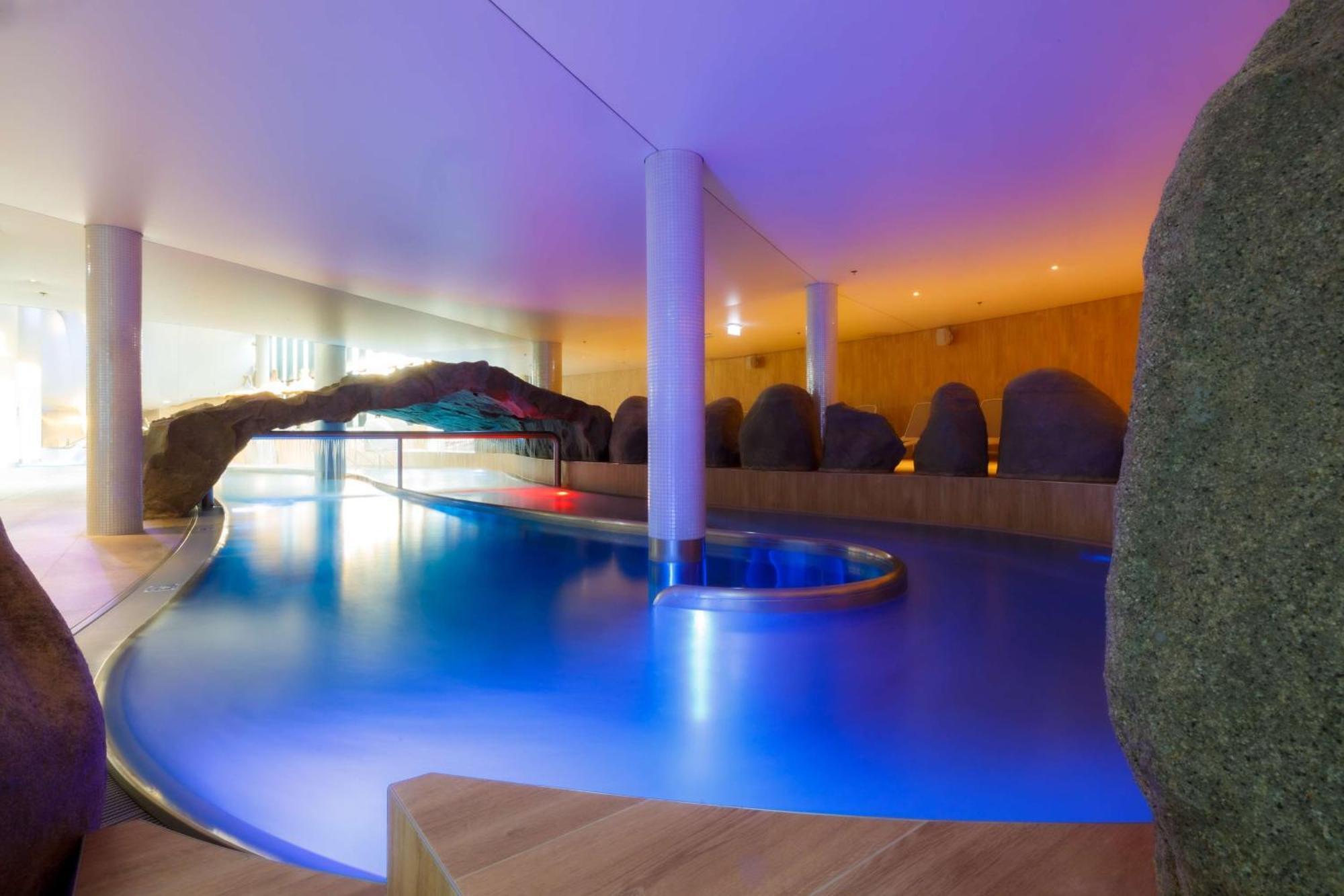 Radisson Blu Resort Swinoujscie Kültér fotó The photo showcases an elegant indoor swimming pool area. The pool has a flowing, organic shape and is illuminated with soft blue and purple lighting, creating a calming ambiance. Surrounding the pool are smooth, natural-looking stones and wooden ele