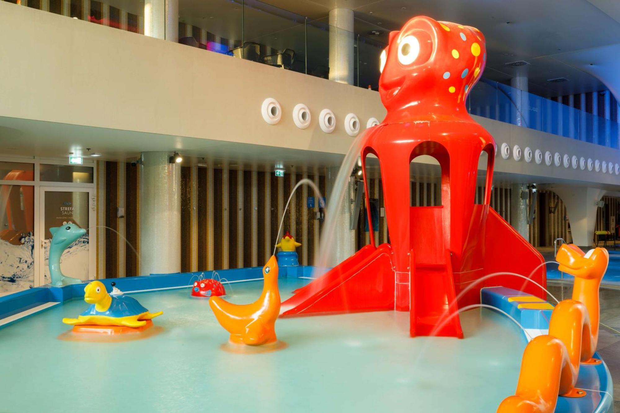 Radisson Blu Resort Swinoujscie Kültér fotó The photo shows a colorful indoor splash pad or water play area designed for children. Central to the scene is a large, bright red octopus structure, which has water fountains spraying out. Surrounding it are various playful, colorful water animals, 